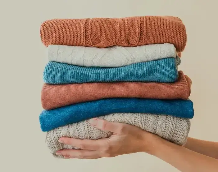 A hand holds a neatly stacked pile of colorful folded sweaters against a plain beige background.