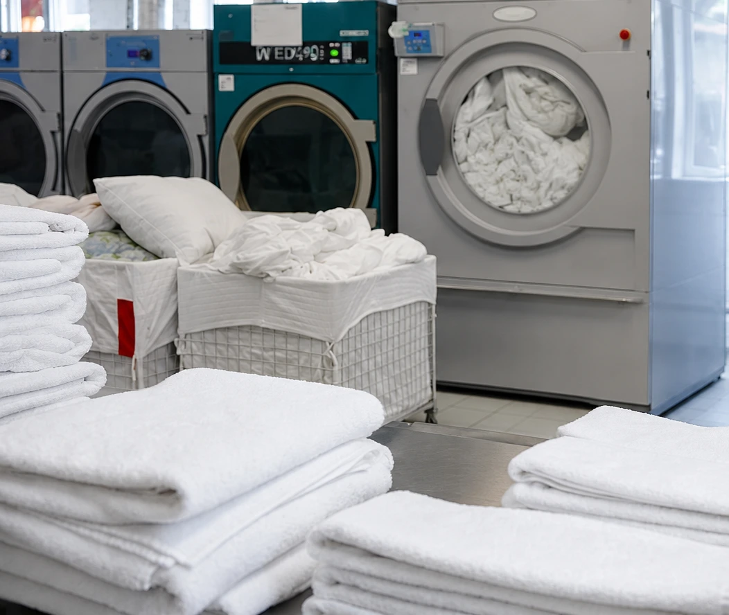 Stacks of folded white towels on a table, with a laundry cart holding linens and large industrial washing machines in the background.