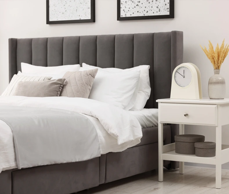 A neatly made bed with white linens and gray headboard sits next to a white nightstand holding a clock, vase with dried grasses, and two round boxes.