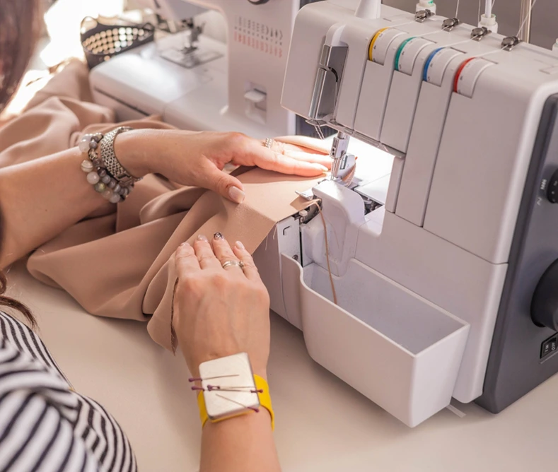 Person using a serger sewing machine to stitch beige fabric, with hands guiding the material and sewing tools visible on their wrist.