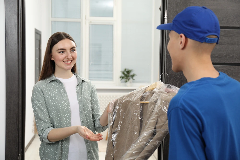 A woman receives a freshly cleaned garment covered in plastic from a delivery person at her door.