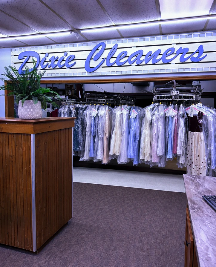 A row of clothes in plastic covers hangs beneath a "Dixie Cleaners" sign inside a dry cleaning shop.