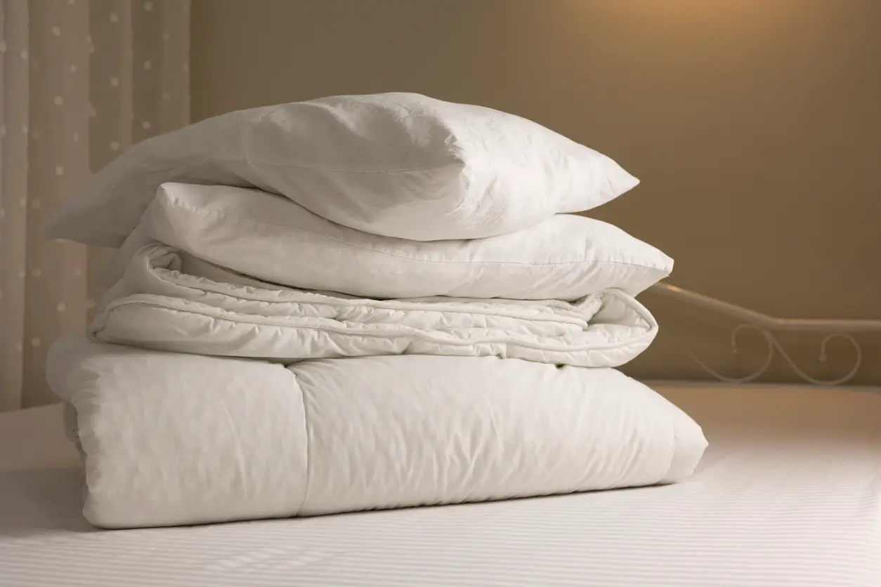 A neatly folded stack of white bedding, including a pillow and comforter, sits on a bed in a softly lit room.