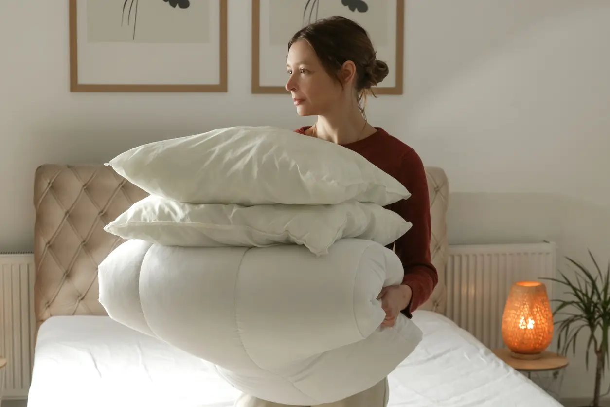 A woman stands in a bedroom holding two white pillows and a folded white comforter, with a made bed and a lit lamp in the background.