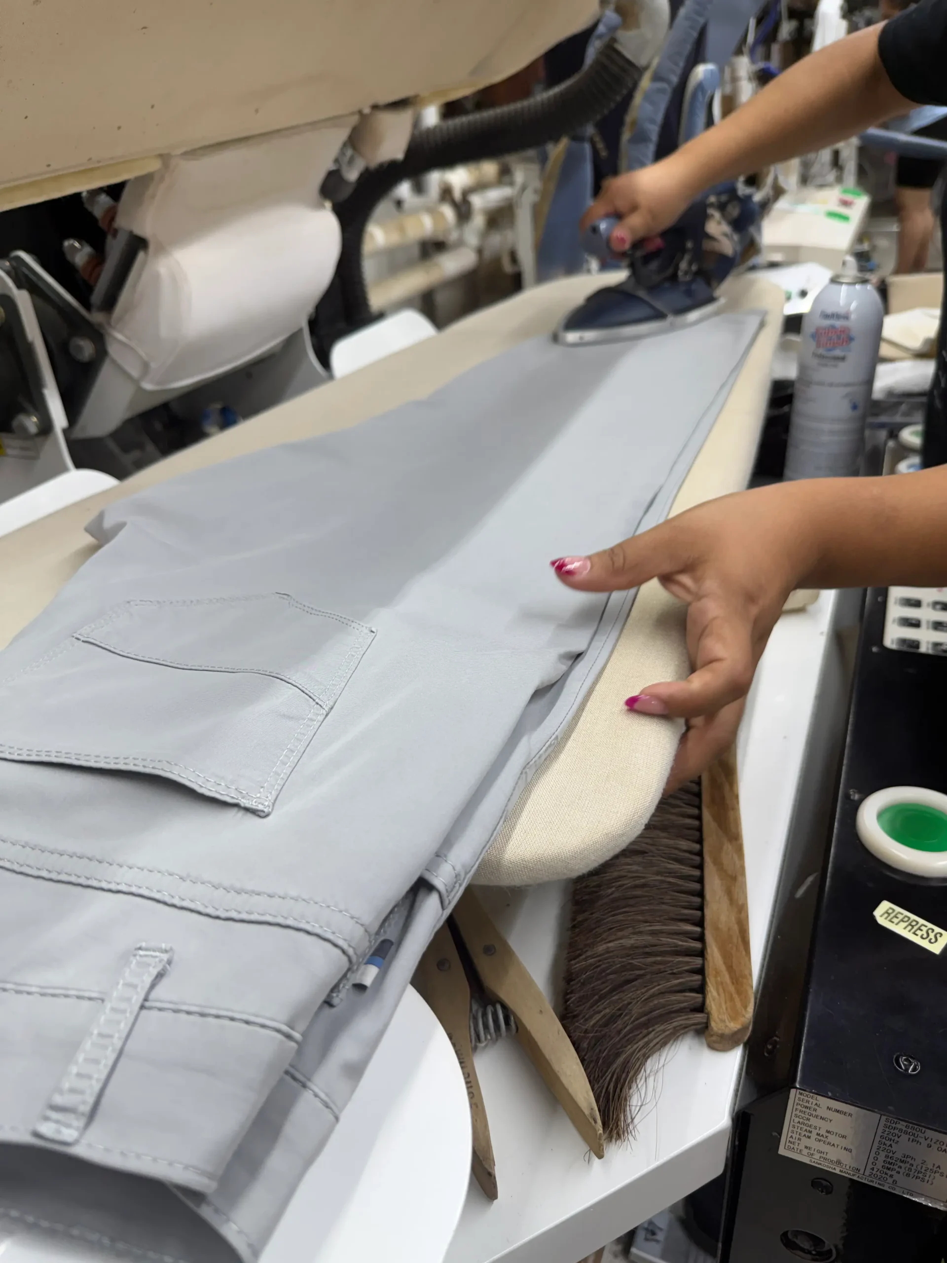 A person presses light gray pants with an iron on an ironing board, surrounded by various laundry tools and supplies.