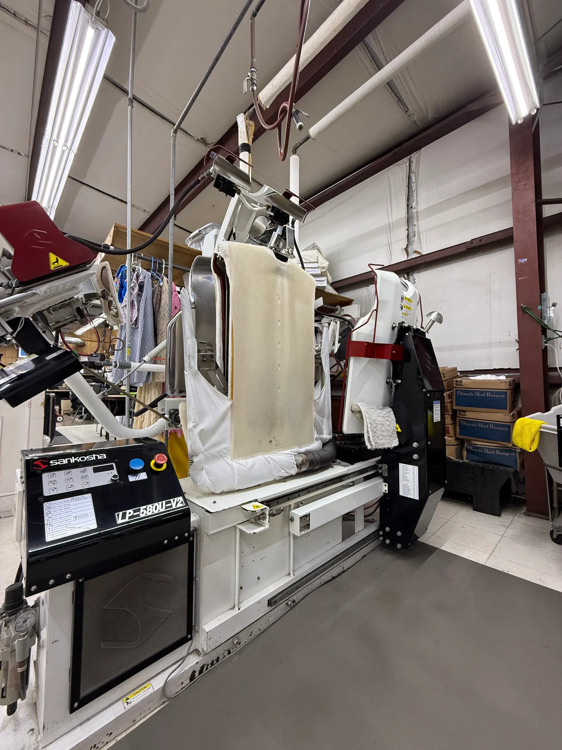 A commercial pressing machine is in a dry cleaning facility, with clothing racks and storage boxes visible in the background.