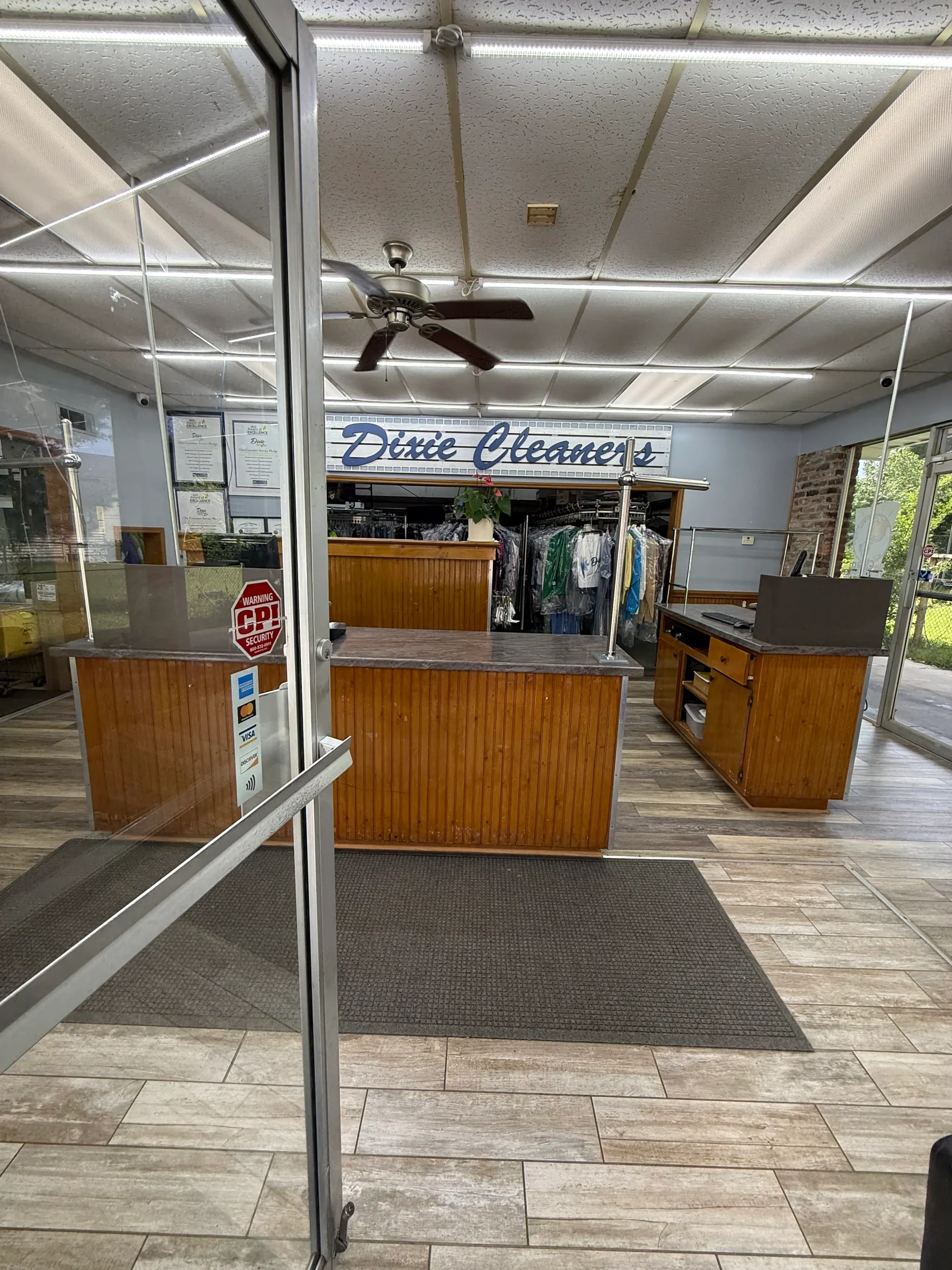 Interior of a dry cleaning shop with wooden counters, clothing racks in the back, tiled floor, and a glass entrance door.