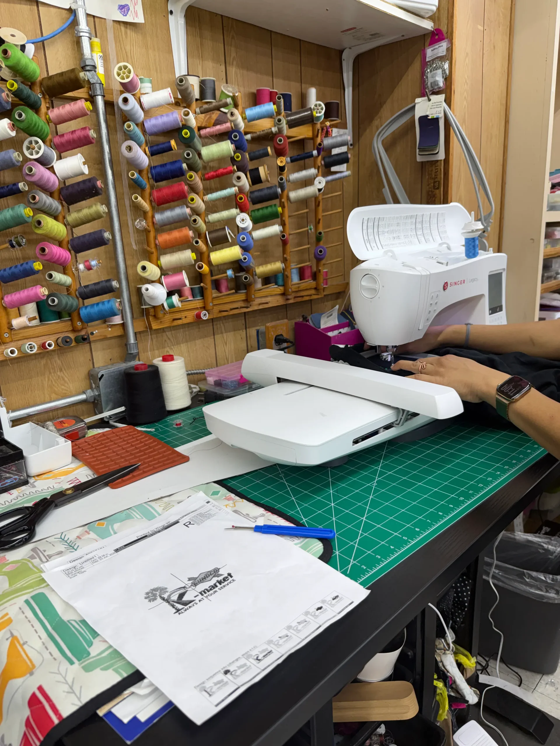 Person operating an embroidery machine on a worktable with thread spools organized on the wall and sewing supplies scattered around.