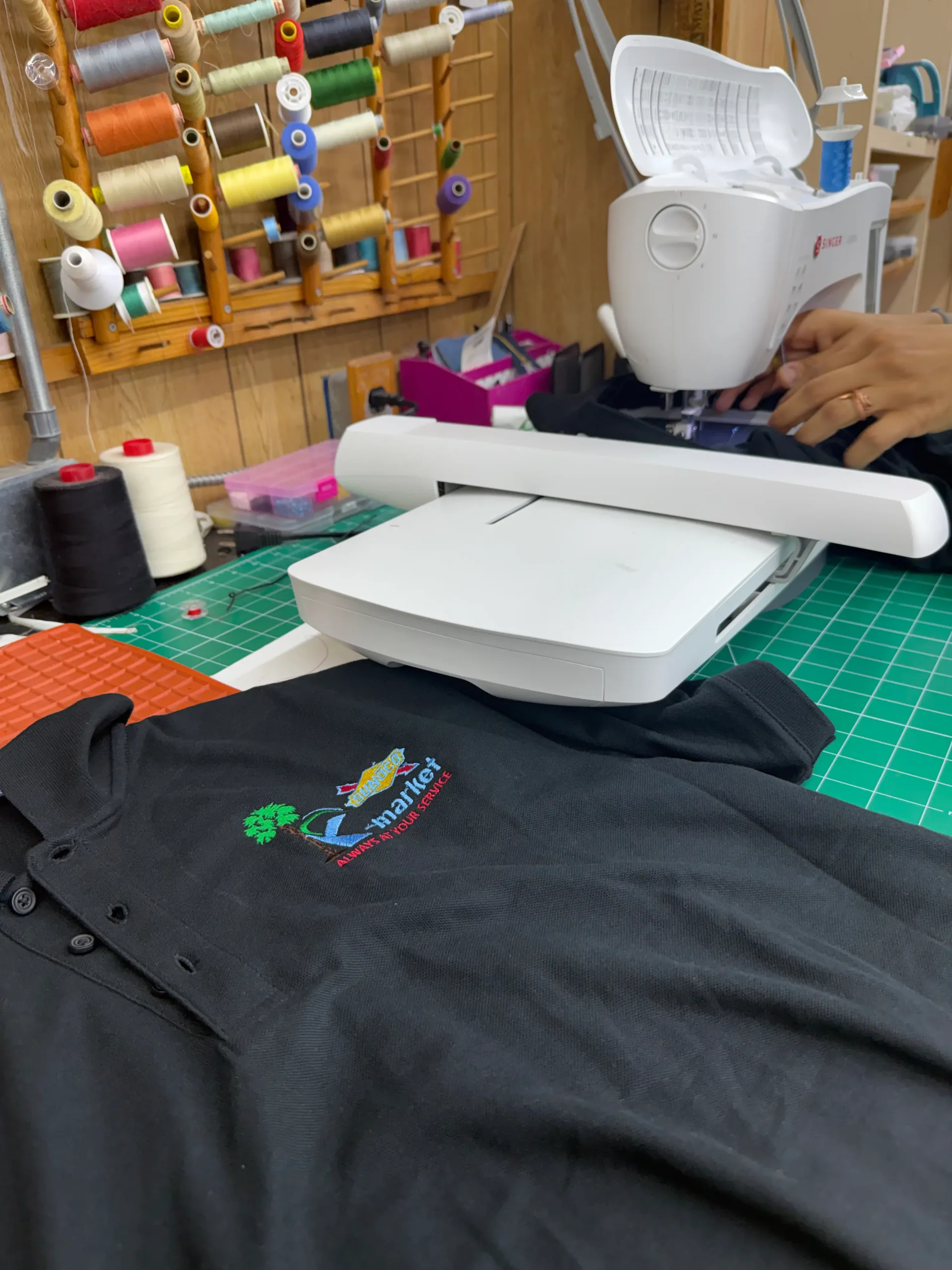 A black polo shirt with colorful embroidery lies on a worktable near a sewing machine, with spools of thread and a person’s hands visible in the background.